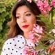 A brown haired woman wearing a white and black
             polka-dotted shirt with a flowery garden in the background.